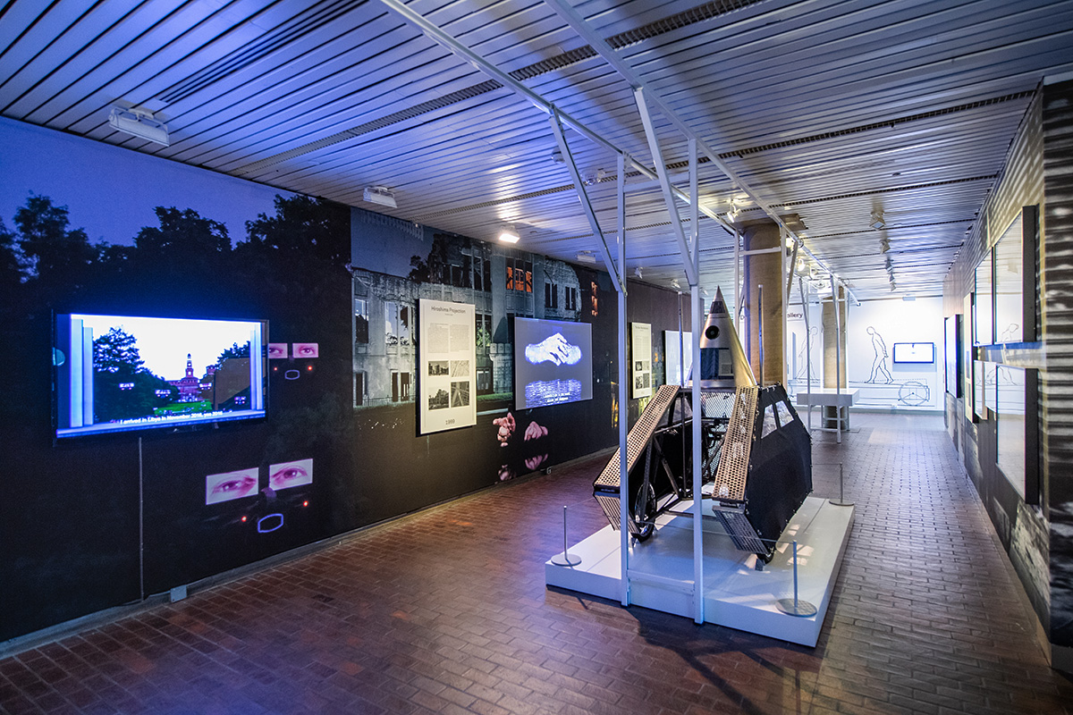 The lobby of Gund Hall with the Interrogative Design exhibit, including wall murals, display cases, projection screens, and Wodiczko’s “Poliscar” vehicle.