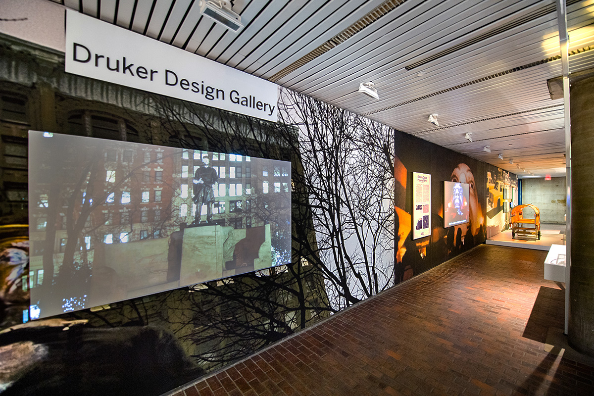 The lobby of Gund Hall with the Interrogative Design exhibit, including wall murals, display cases, and projection screens.