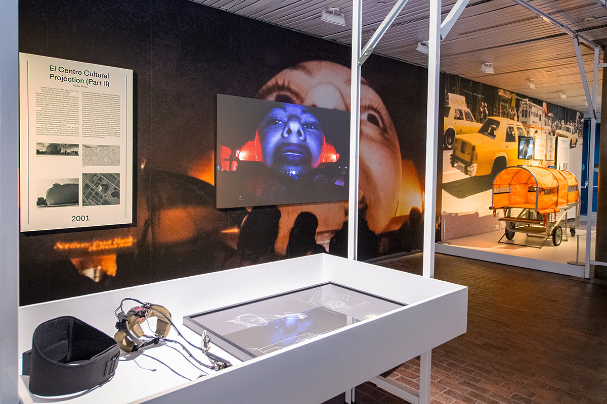 The lobby of Gund Hall with the Interrogative Design exhibit, including wall murals, display cases, and projection screens.