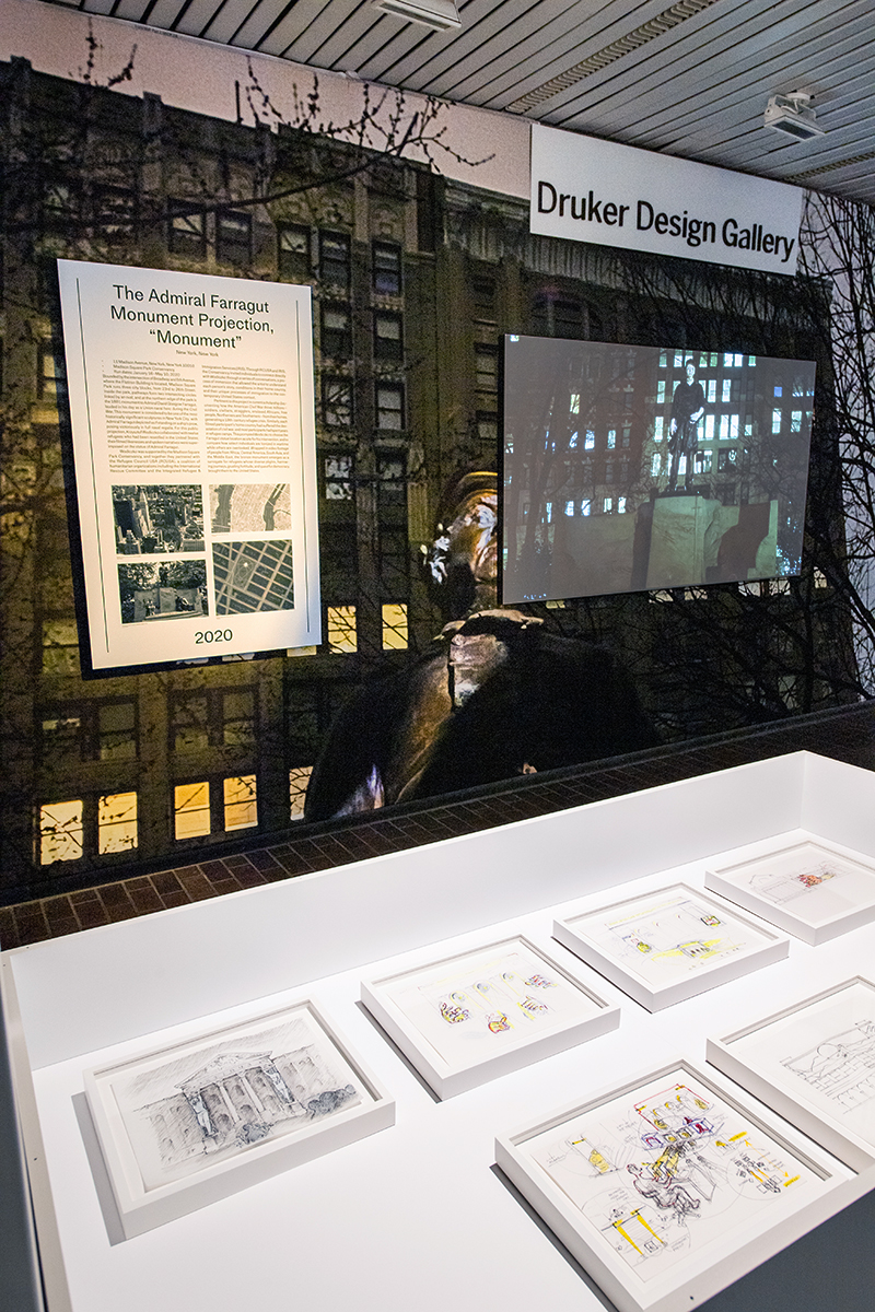 The Interrogative Design exhibit in Gund Hall, with a display case of Wodiczko’s sketches in front of a wall mural and projection screen.