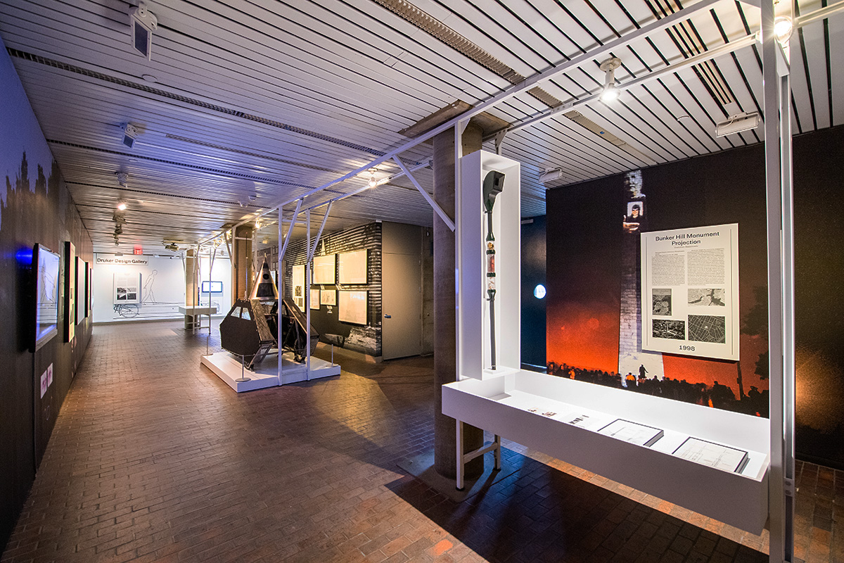 The lobby of Gund Hall with the Interrogative Design exhibit, including wall murals, display cases, projection screens, and Wodiczko’s “Poliscar” vehicle and “Alien Staff”.