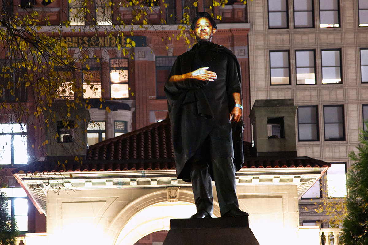 A monument of Abraham Lincoln at night with city buildings in the background. A person’s face and hands are projected onto the face and hands of the Lincoln statue.