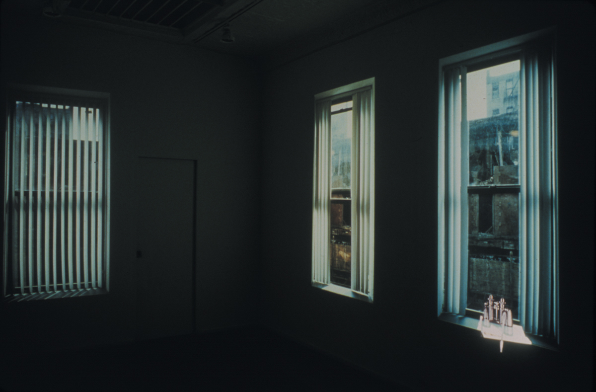 A dark empty room with bare white walls and a plain door. On the walls are three projections of different windows with views of New York City buildings. A pair of binoculars sits on one of the window sills.