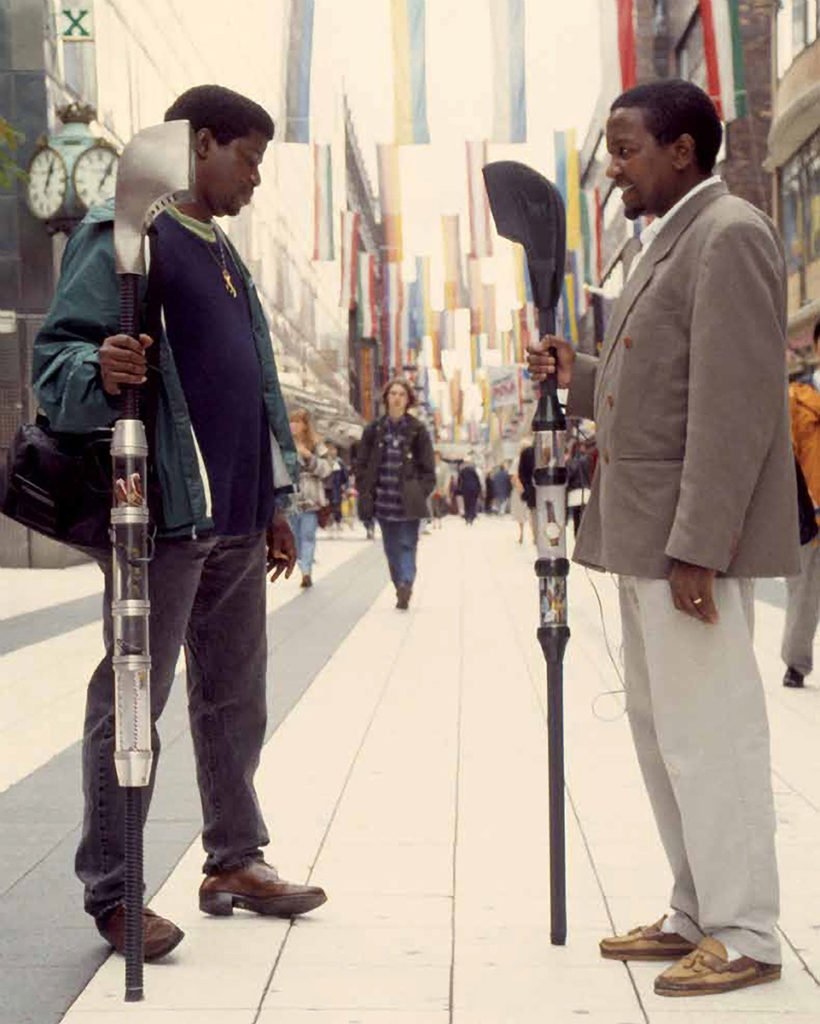 A busy pedestrian area between city buildings. Two people stand facing each other, each holding a unique version of the Alien Staff which they hold upright to face the other person.