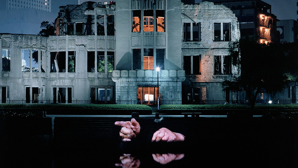 The facade of the Hiroshima Peace Memorial at night, a stone ruin of a city building on the edge of a river. Two hands are projected on a stone wall above the surface of the water and reflected in the water below.