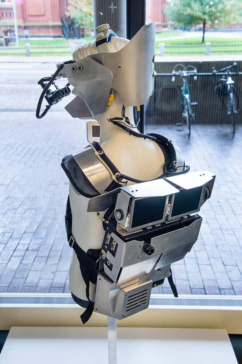 Back view of a mannequin displaying the Dis-Armor, an apparatus made of metal and electronics worn over the torso and head.