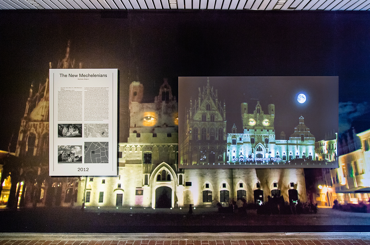 A wall in Gund Hall showing a mural, with text panel and projection screen on top.