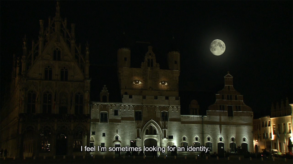 The medieval-era city hall building in Mechelen, Belgium at night, with a projection of a person’s eyes on the tower. At the bottom a subtitle reads, “I feel I’m sometimes looking for an identity.”