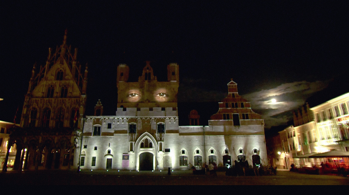 The medieval-era city hall building in Mechelen, Belgium at night, with a projection of a person’s eyes on the tower.