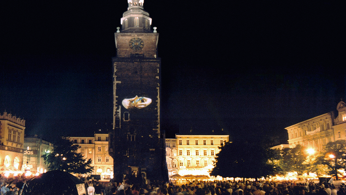 A stone clock tower in the middle of a crowded town square at night. On the face of the tower is a projected image of two hands peeling a potato with a knife.