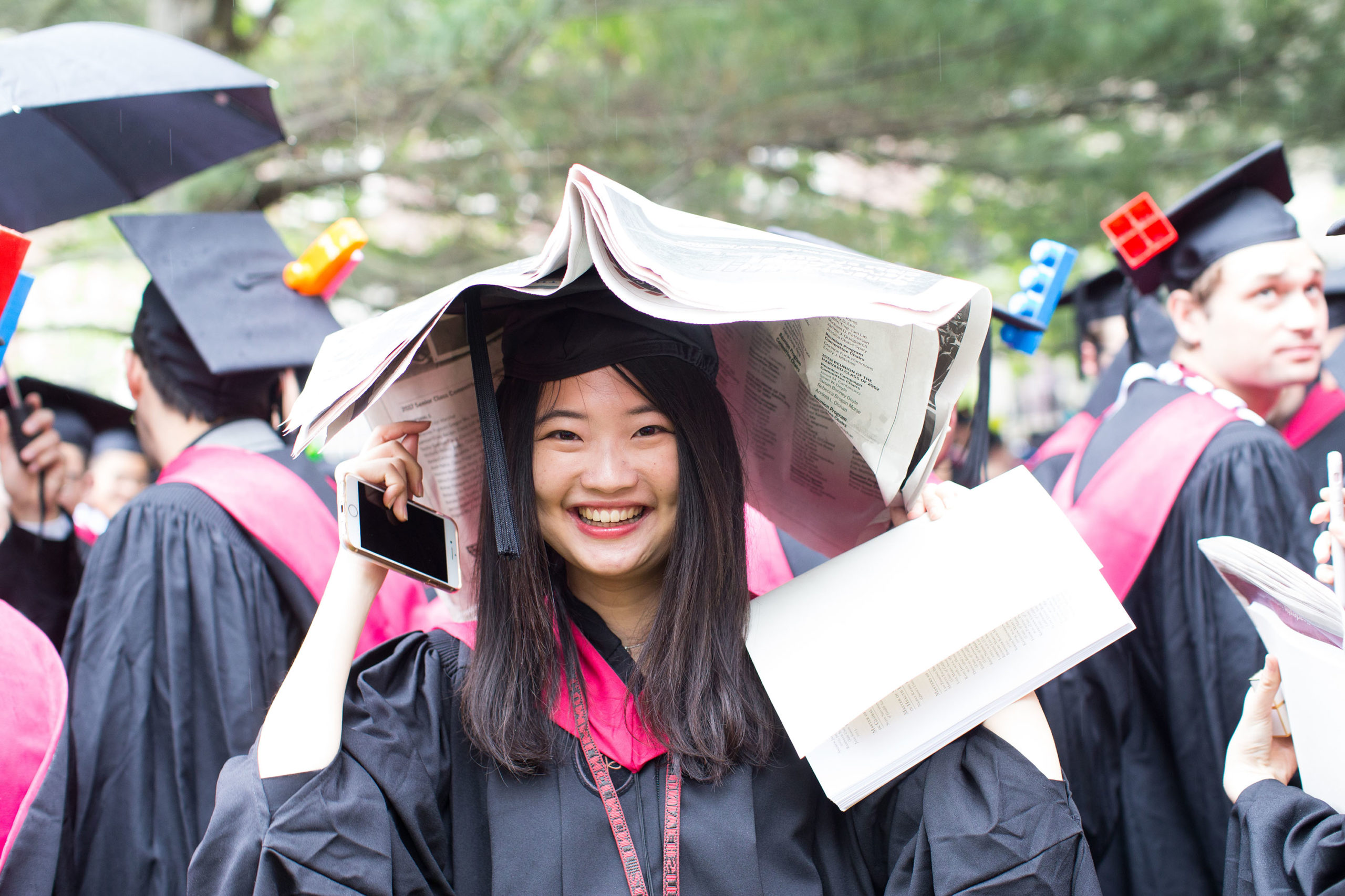 Woman dressed in graduation gown smiles at the camera while holding a newspaper above her head to shield her from the rain.
