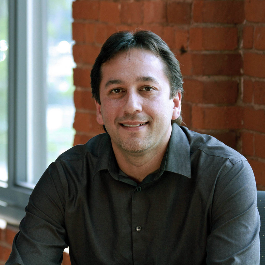 Headshot of Sam Olbekson, who sits in front of a brick wall and wears a gray collared shirt.