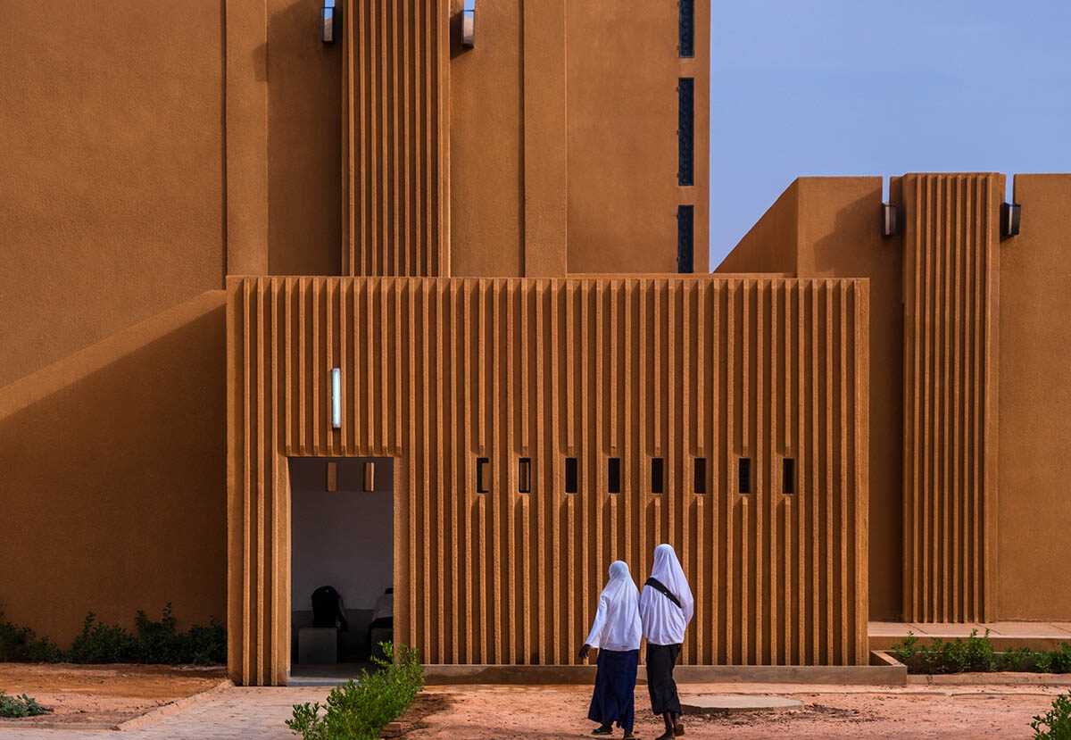 Two figures stand in front of a tall brown building with multiple tiers.