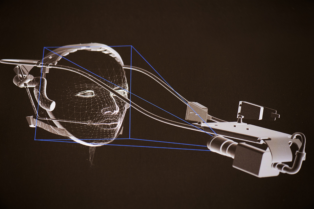 CAD drawing of a person’s head wearing an apparatus that extends forward and holds a video camera pointed back towards the face.