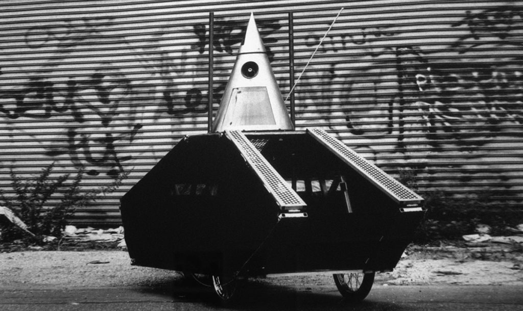 Black and white photo of a polygonal-shaped vehicle built on three bicycle wheels, with an inverted cone shape on top made of metal with a clear window on the front.