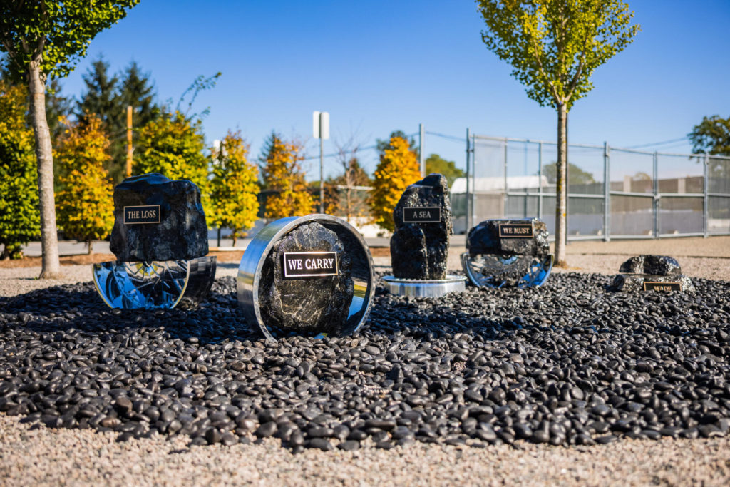 Outdoor sculpture made up of boulders and metal. The words "The loss we carry a sea we must wade" are attached to the boulders.