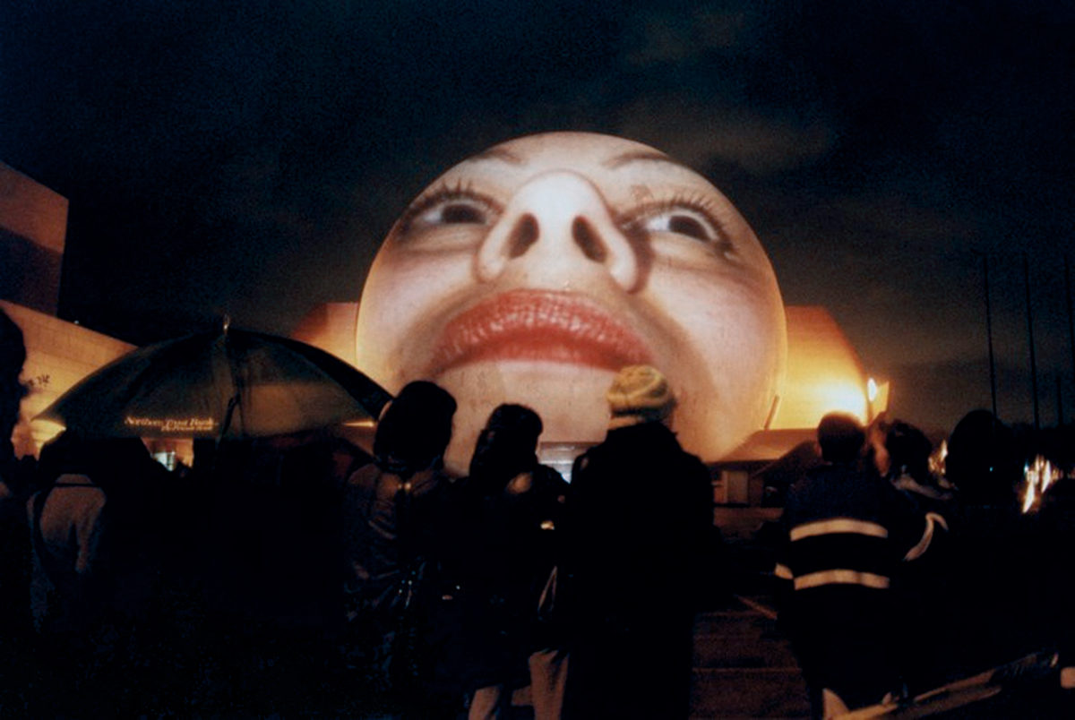 Art installation with a large face projected on an orb, outdoors during the nightime.