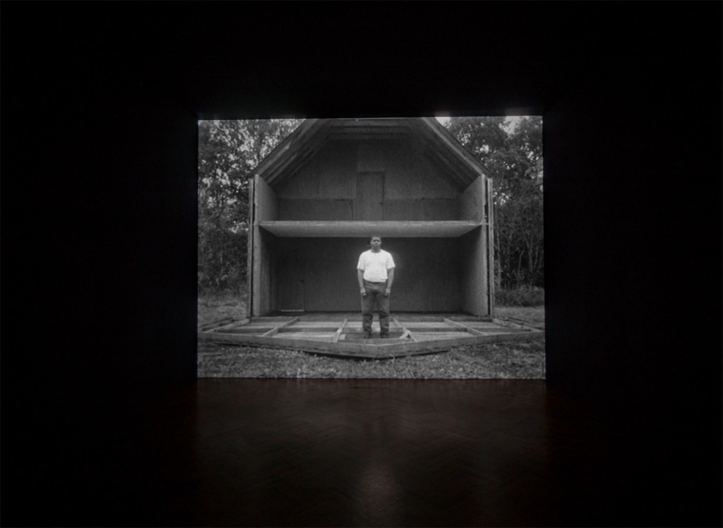 Black and white image of man standing in white shirt in the middle of a fallen house façade; interior of empty house pictured in background.
