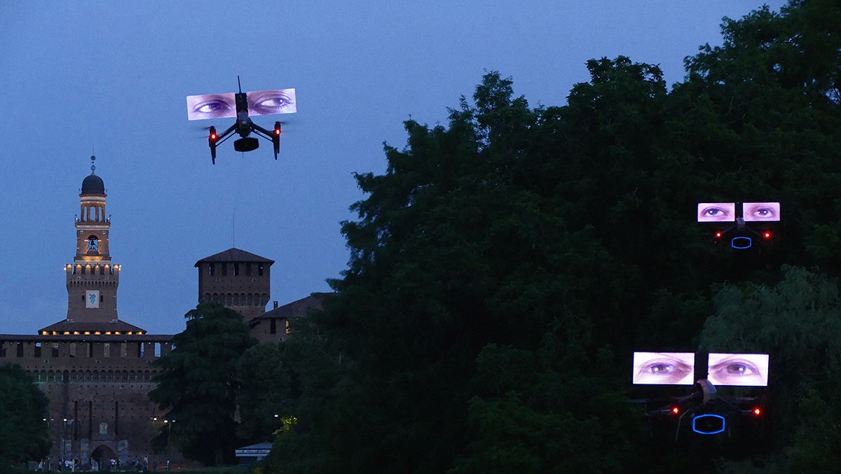 An outdoor park area at dusk, with trees and a medieval structure in the background. Three drones carrying video screens of human eyes hover in the air.