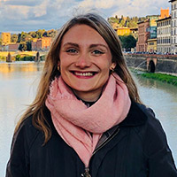 Headshot of Livesey Pack. Livesey has long blond hair and wears a black jacket and large pink scarf in front of a city landscape.