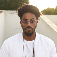 Headshot of Muha Bazila. Muha has curly dark hair and a beard, Muha wears thin-rimmed glasses, a white shirt, and bomber jacket. 