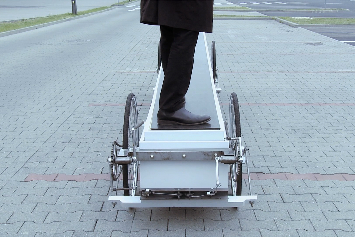 A man stands at one end of a long low vehicle made of two long boxes with bicycle wheels and drivetrain attached.