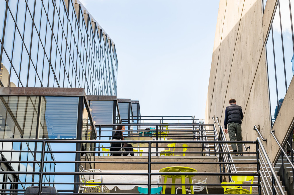 A photo of the balconies on each level of Gund Hall. There is a person walking up the stairs that connect each balcony level, and a person sitting on a colourful table, working on their laptop.