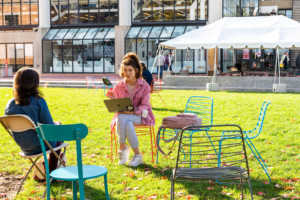 A photo of students sitting in the GSD backyard, a grassy area between Gund Hall and adjacent GSD buildings, populated with colourful chairs.