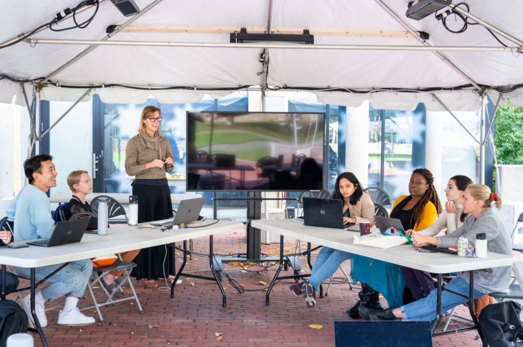 An image of an instructor teaching a class outside under a tent in the GSD backyard. There is a screen that has been set up, and the student are sat at tables.