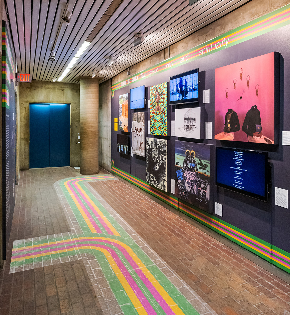 A view of the exhibit in Gund Hall showing a variety of colorful artwork on the walls and colorful stripes decorating the floor.