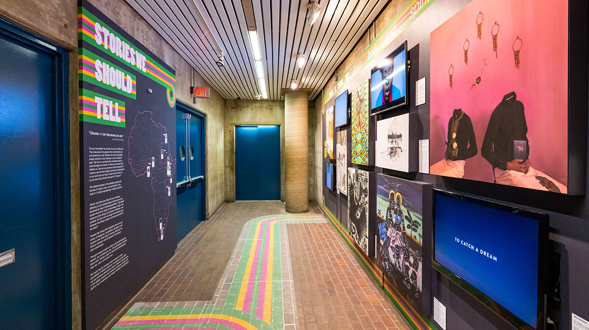 A view of the exhibit in Gund Hall showing a variety of colorful artwork on the right-hand wall, a map of Africa on the left-hand wall, and colorful stripes decorating the floor.