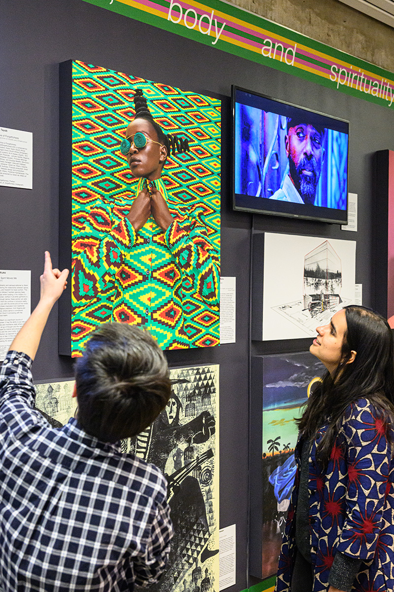 Two people looking at a colorful portrait on a wall with multiple pieces of artwork and video screens.