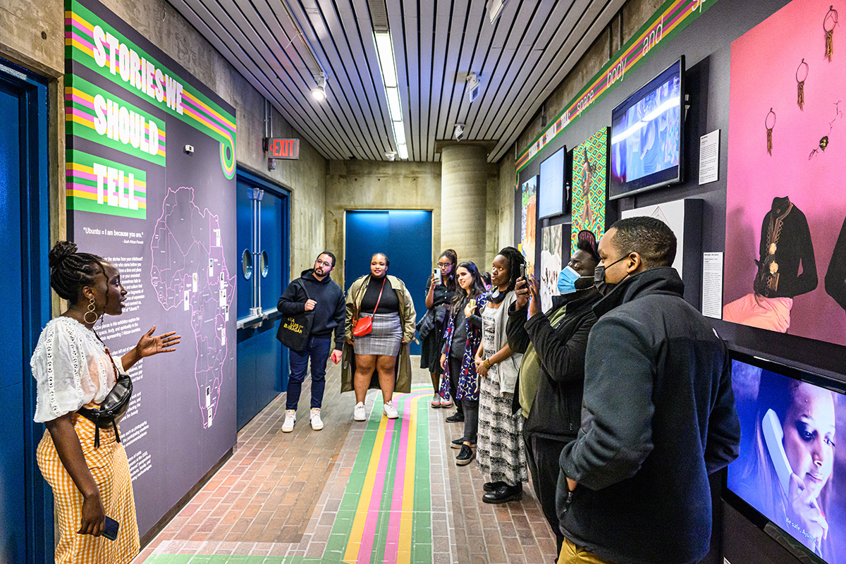A person talking to a group of listeners in front of an exhibit of multiple colorful pieces of art and a map of Africa.