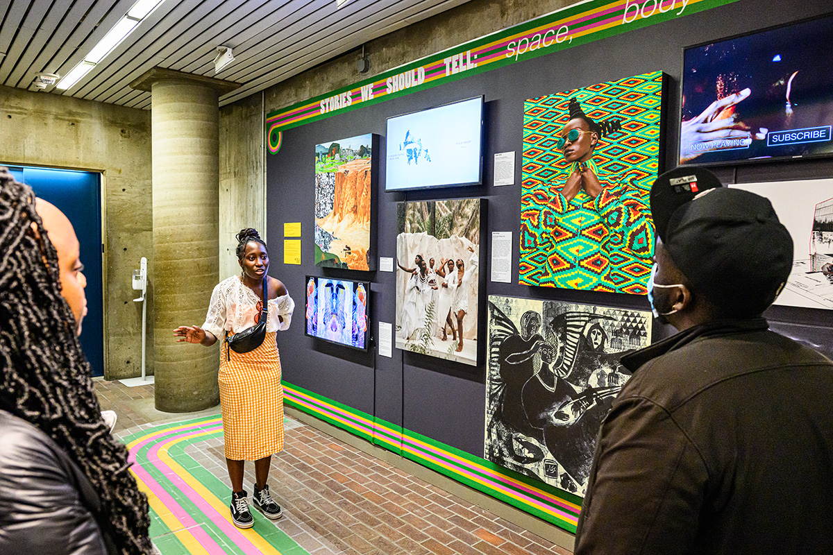 A person talking to a pair of listeners in front of a wall with multiple colorful pieces of art.