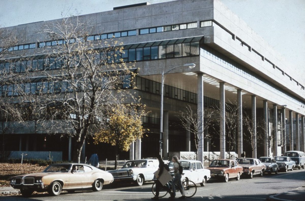 Archival photo of Gund Hall from Quincy Street with cars parked out front and students walking a bicycle across the street.