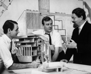 Black and white photo of three architects talking around a model and drawings.