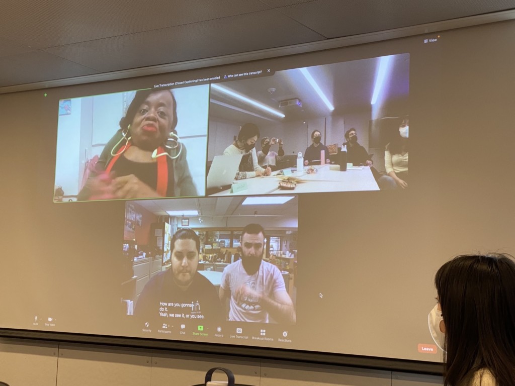 A projector screen displaying a zoom meeting where students discuss adaptive design with speakers who are joining online.