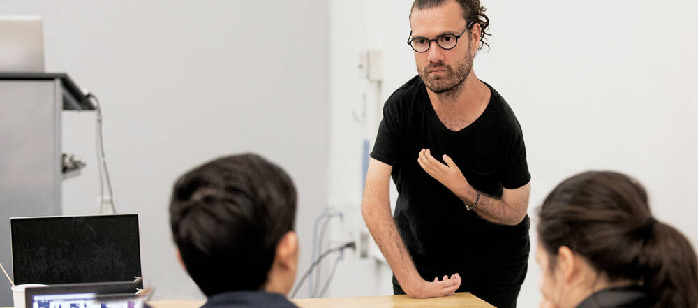 Designer Josh Halstead leans on a desk, speaking to students.