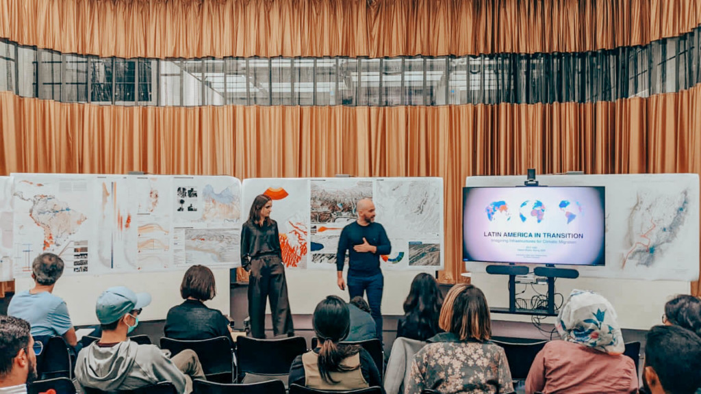 Instructors present a powerpoint in front of the piper auditorium gold curtain and drawings pinned to a board.