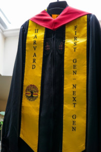 First Gen Next Gen stole hangs on a hanger with a graduation robe. The stole is yellow and features a drawing of a tree with long roots and flowers and the words Next Gen.