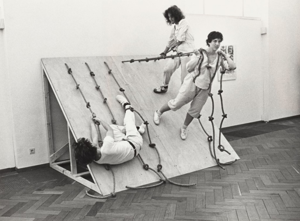 Dancers performing on inclined wood board with ropes.