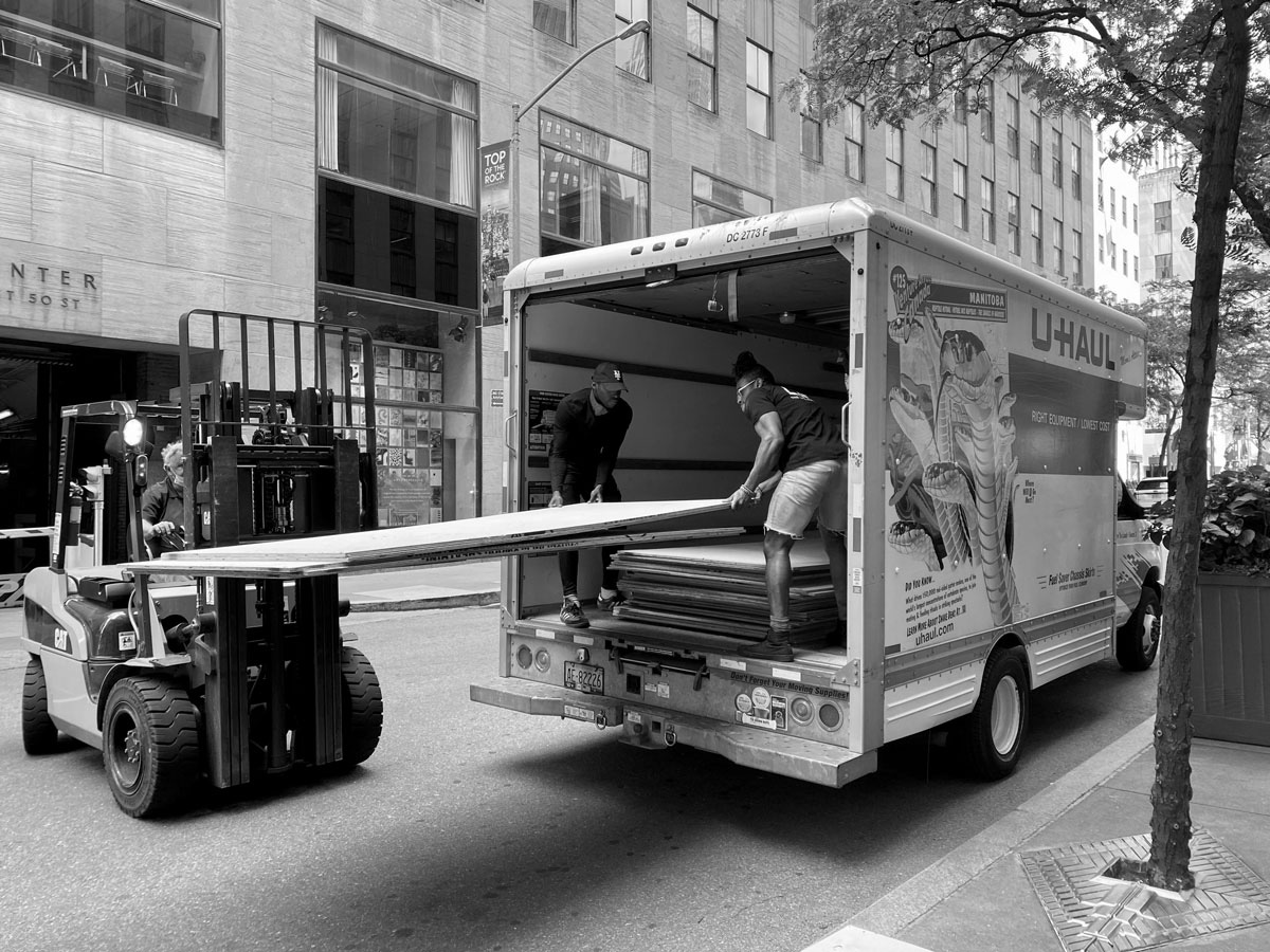 Two people guide a large sheet of plywood onto a van with the help from a fork lift.