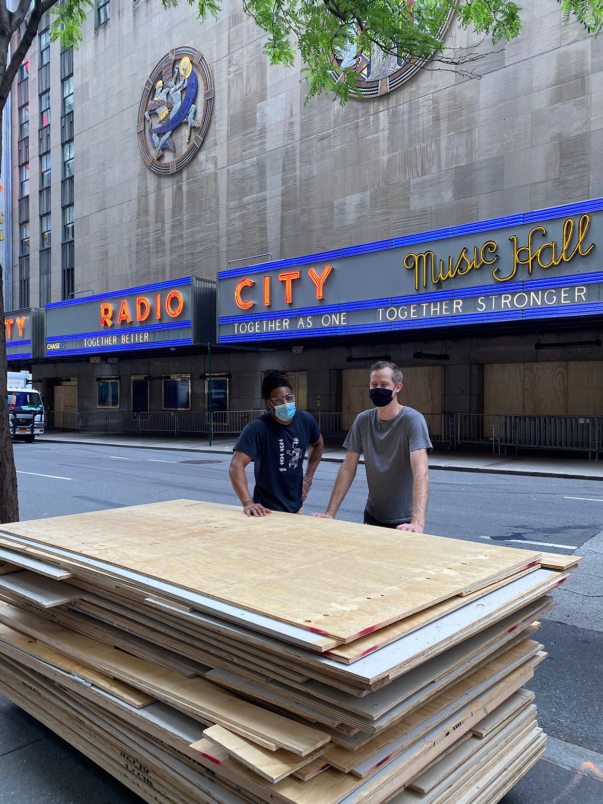 Two people lean on a tall stack of plywood with happy expressions.