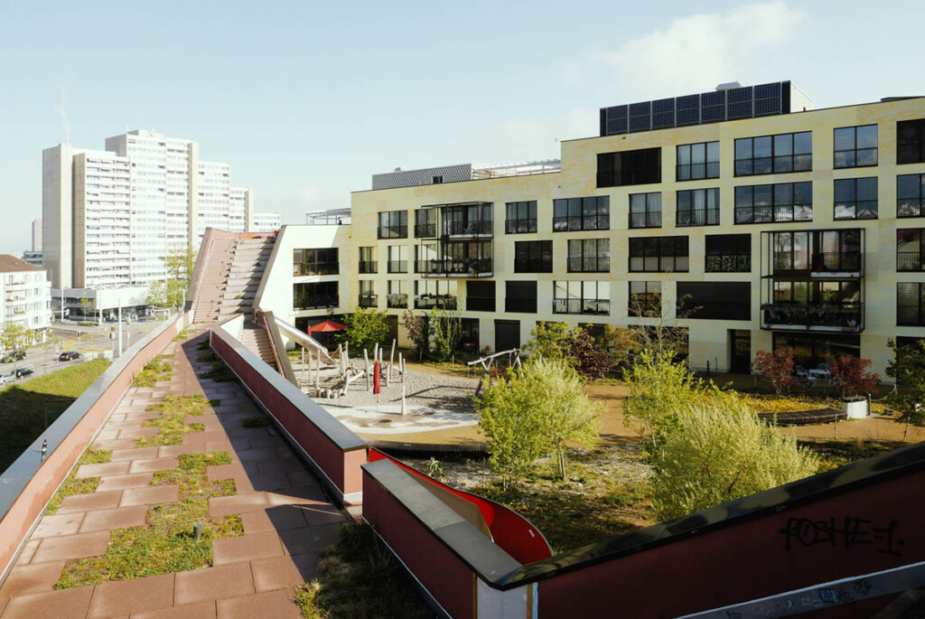 A walkway leads to a building complex with many windows and some trees in a plaza.