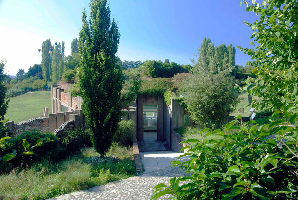 SITE Rossini Pavilion in Briosco, Italy. A entrance to a building is surrounded by lush green plants and trees.