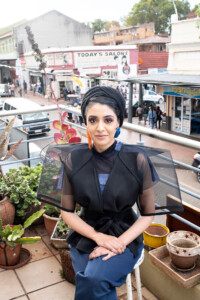 Headshot of Sumayya Vally, who sits on a balcony above a busy street.