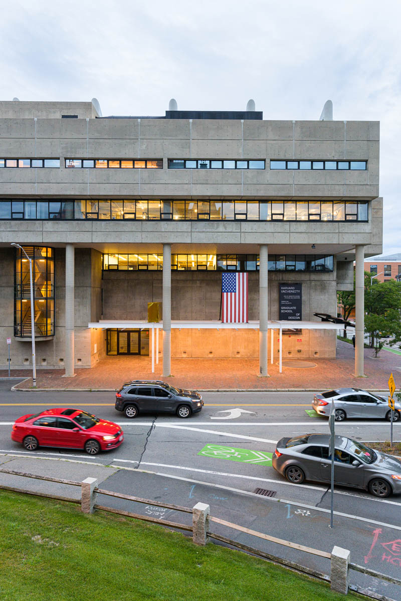 Exterior of Gund Hall in the evening with the paviolion lighting up the sidewalk. Cars are moving on the road infront of it.