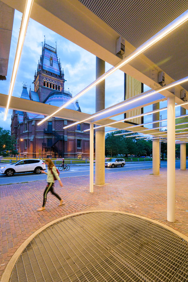 Person, lit by thin lighting strips, walking through the pavilion's colum next to a large circular grate; the street and Memorial hall is in the background.