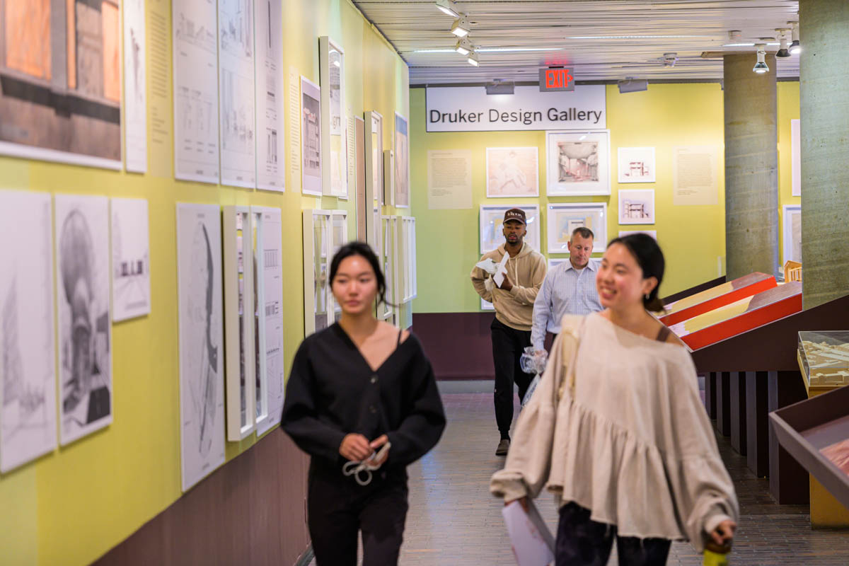 People looking at and traversing through the exhibition corridor. On the walls are framed drawings, and reprodcutions. Down the middle of the corridor is a canted display case showing models and archival artifacts.