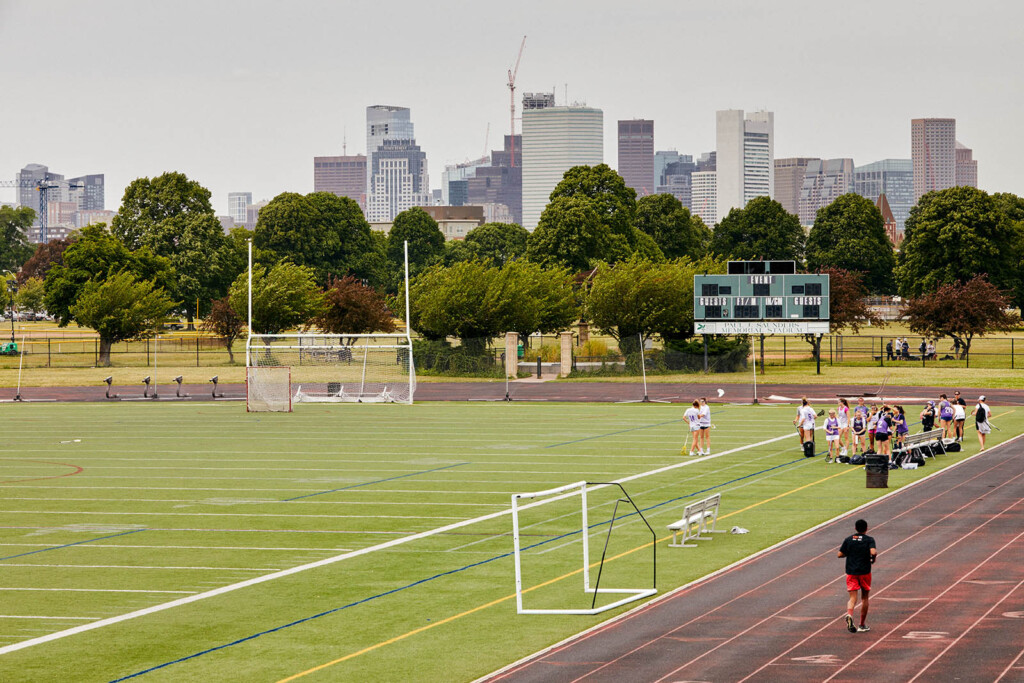 A close-up of a track with a man running and a group of people gettrin ready to exercise. Skyline of a city in a background.